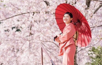 桜の花を背景に赤色の着物姿で立つ女性の画像